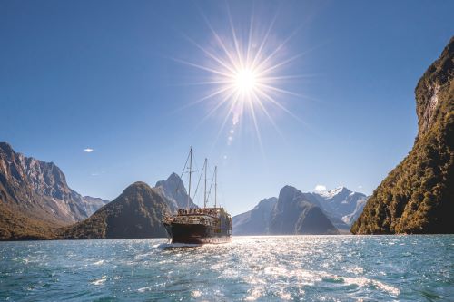 A boat sails on the tranquil, sunlit waters of Milford Sound, surrounded by towering mountains under a bright blue sky.