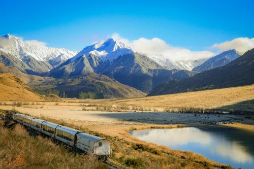 A scenic train journey passes through a mountainous landscape with snow-capped peaks and a clear blue sky.