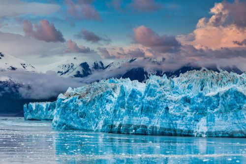 Massive blue glacier with jagged ice formations, calm reflective water in the foreground, and snow-covered mountains under a colorful sky in the background.