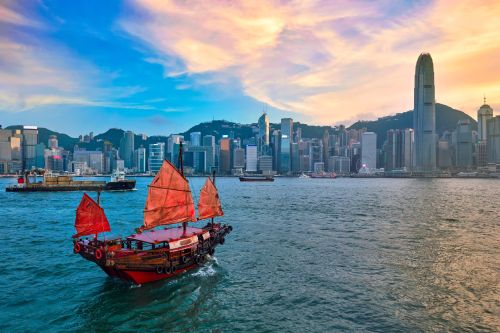 A small boat with red sails in the water in front of a city landscape at dusk