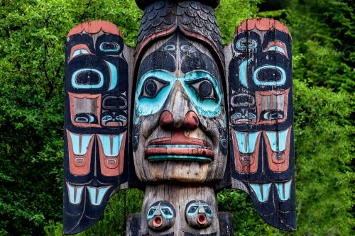 Colorful carved totem pole with intricate designs set against a backdrop of green foliage.