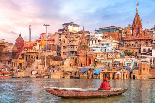 Man on a small wooden boat riding along the Ganges River with Varanasi city in the background.