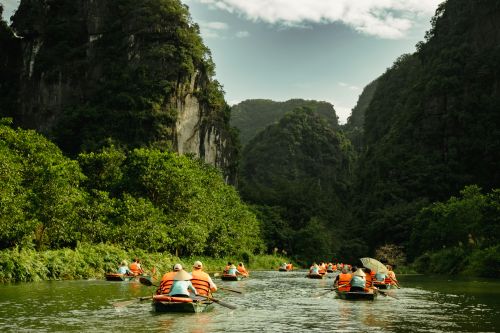 Several small boats carrying people in orange life jackets travel along a calm green river, surrounded by steep limestone cliffs and dense tropical vegetation under a partly cloudy sky.