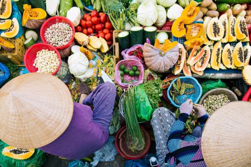Overhead view of a busy market scene with two people wearing conical hats sorting and selling a variety of colorful fresh produce, including papaya, pumpkins, tomatoes, beans, and leafy greens, arranged in bowls and baskets on the ground.