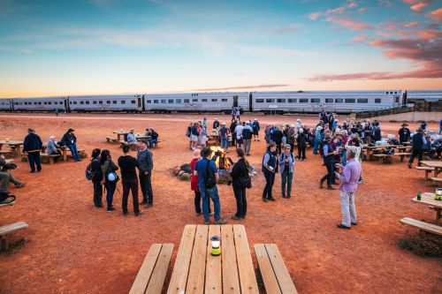 JGroup of people around picnic tables and a campfire near the parked train in the red-soil outback at sunset.