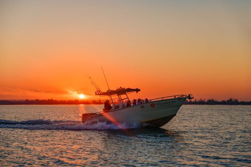 A fishing vessel cruising with the sun setting behind it and colouring the sky in orange. 