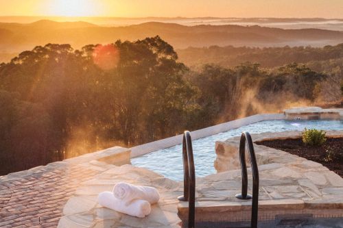 Steam rising from a stone-lined heated pool overlooking forested hills at sunrise, with white towels nearby.