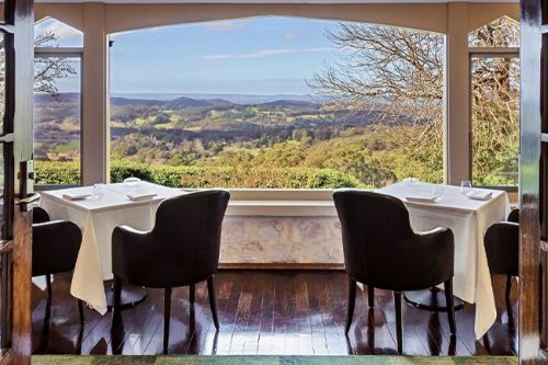 Two tables set for dining beside large windows overlooking green hills and open countryside.