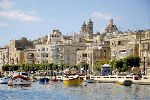 Colourful boats moored in a Mediterranean harbour with a historic city skyline behind.
