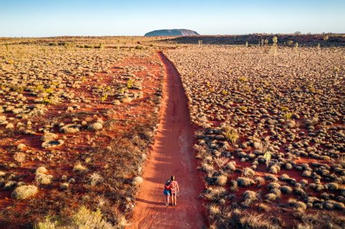 A couple standing in front of a long red dirt road leading to Uluru in the background 