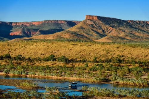 A vehicle crosses a river in front of dry grasslands and flat-topped red mountains.