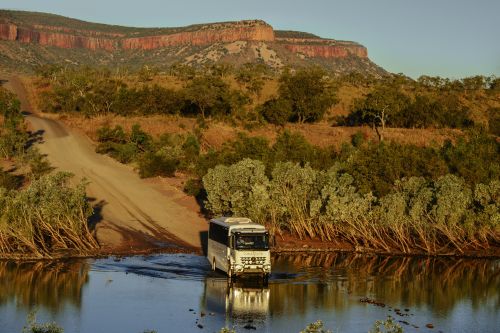 Tour bus drives through water near a mesa in a remote, sunlit landscape. 