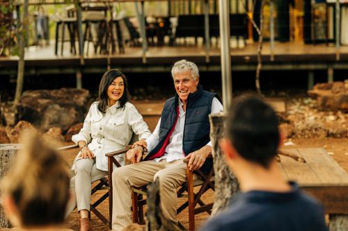 Two people sit and chat in wooden chairs at a rustic outdoor lodge with others nearby.