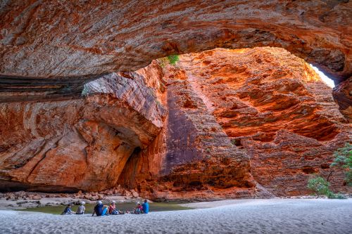 A group of people sit on a sandy riverbank beneath a massive natural rock arch in a red sandstone gorge, with layered rock walls glowing orange in the reflected sunlight and a shallow river flowing nearby.