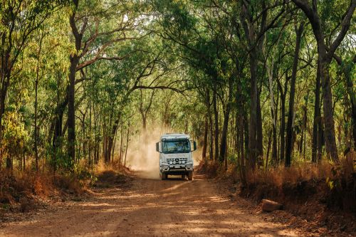 A four‑wheel‑drive coach travels along a dusty, unsealed road through tall eucalyptus woodland, with sunlight filtering through the trees and a cloud of red dust trailing behind the vehicle.