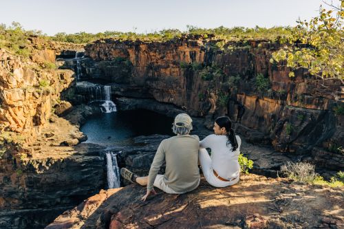 Two people sit on a rocky ledge overlooking a deep sandstone gorge with cascading waterfalls and a clear pool below, surrounded by rugged cliffs and sparse vegetation under bright daylight.