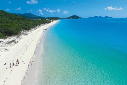 Aerial view of a curved white-sand beach with turquoise water and dense greenery.