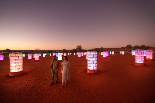 Colourful cylindrical lights scattered across a desert at dusk, with two people walking nearby.