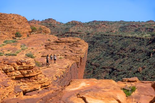 Four people stand on a rocky cliff edge overlooking a deep canyon in a desert landscape.