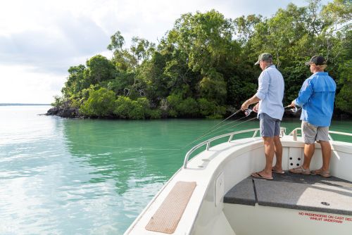 Two people fish from a small white boat in calm turquoise water near a forested shoreline.