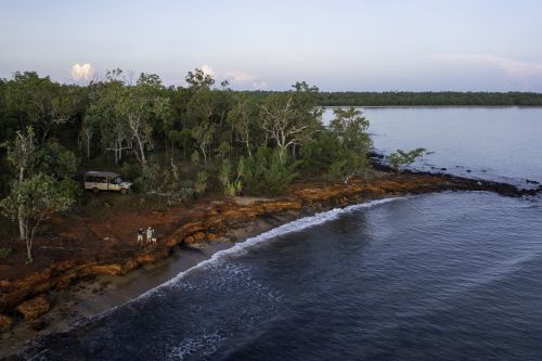 People stand near a parked vehicle on a rugged beach bordered by dense forest.