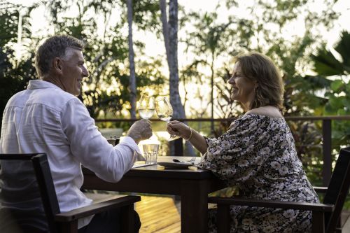 Two people clink wine glasses at an outdoor dining table surrounded by tropical greenery.