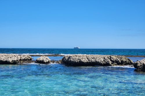 Clear blue ocean with coral formations in foreground and a cruise ship on the horizon under sunny skies.