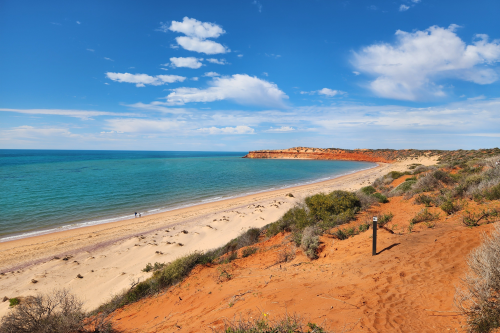 Scenic beach bordered by turquoise ocean and reddish-orange cliffs under a partly cloudy sky.