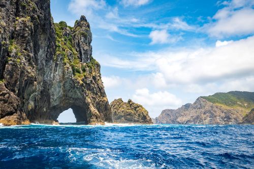 Rock arch and rugged coastline in the Bay of Islands, viewed from the water under a blue sky.