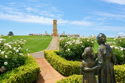 Statue of two people facing a memorial tower surrounded by green lawns and white roses.