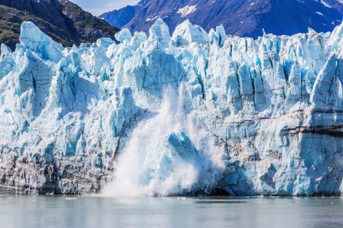 Large chunk of ice calving off a towering blue glacier into calm water with mountains in the background