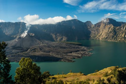 Volcanic crater with a steaming cone beside a turquoise lake surrounded by mountains
