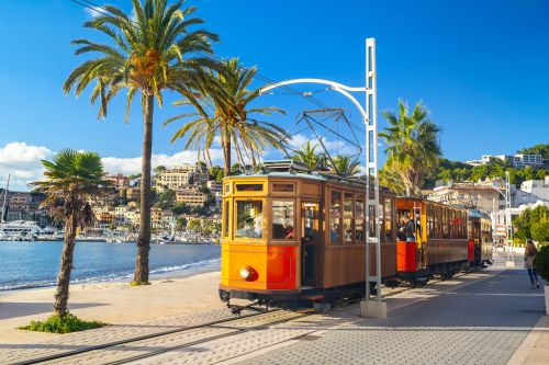 A bright orange tram runs along the waterfront lined with palm trees in Spain