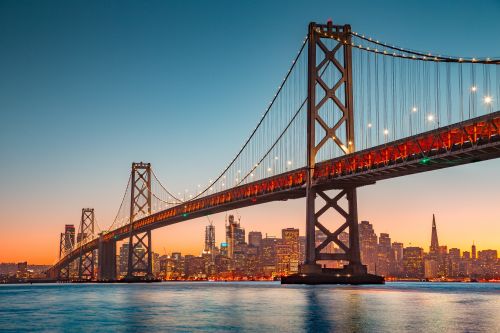 Bay Bridge illuminated at sunset with city skyline in the background and calm water in the foreground