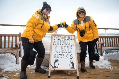 Two travellers in yellow Quark Expedition parkas are celebrating their Antarctic circle crossing by toasting with a drink and smiling in the camera.