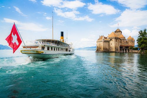 A boat with a Swiss flag at the back heading towards a castle built by the lake