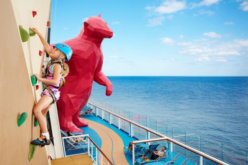 A girl climbing up a rock wall aboard the vessel with their parent relaxing on a lounge.