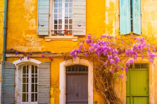 A yellow European building with coloured window covers and purple flowers 