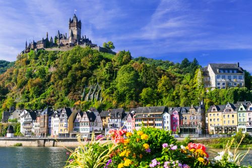 A medieval castle sitting ontop of a hill with with old European houses lining the river underneath