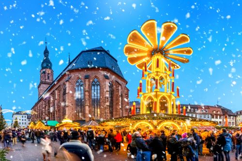Snow falling down on a lit up Christmas market with lots of people mingling around the stands 