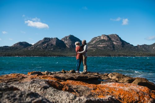 Two people stand on a rocky shoreline, looking across a bright blue bay towards a rugged mountain range under a mostly clear sky.