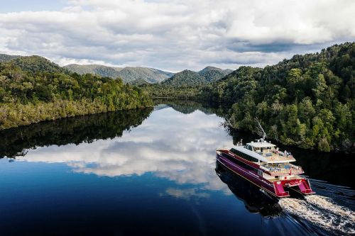 A boat cruises along a calm river surrounded by dense forested hills, with clouds reflected in the still water.