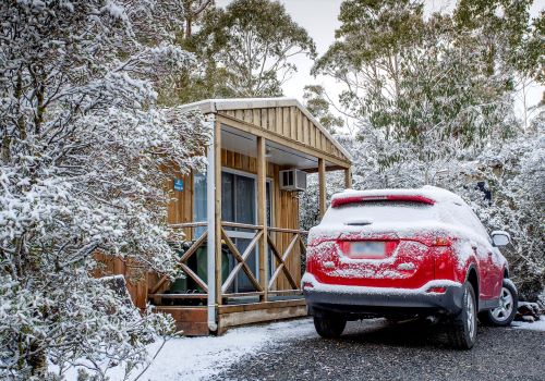 A red car covered in snow is parked in front of a wooden hut in the middle of the snowy forest 