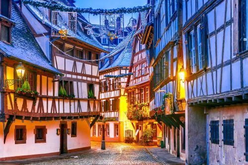 Narrow cobblestone street with traditional half‑timbered buildings, glowing streetlights, and festive decorations overhead in a historic European old town.