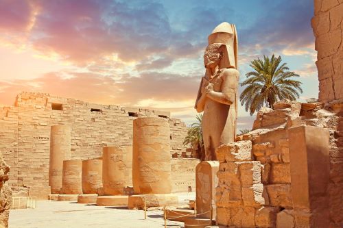 The imposing statue of Ramses II at Karnak, Egypt, illuminated by the warm hues of sunset is showcasing the grandeur and craftsmanship of ancient Egyptian art and architecture.