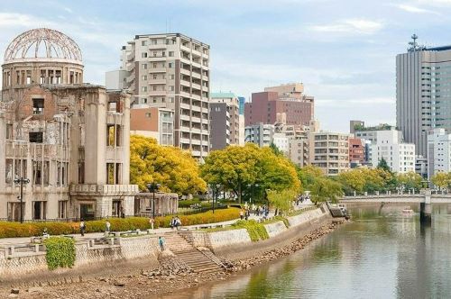 A building lying in ruins besides the riverbed in Hiroshima with modern, intact buildings in the background. 