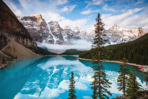 A rocky and snow-covered mountain range is reflecting in the prisine blue water of a still lake which is surrounded by a pine forest