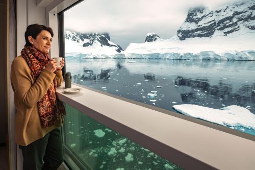 Person holding a cup while looking out a large window at icy waters and snow-covered mountains.