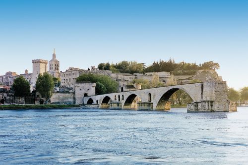 A historic stone bridge crossing a river, with medieval buildings and a palace in Avignon, France, in the background.