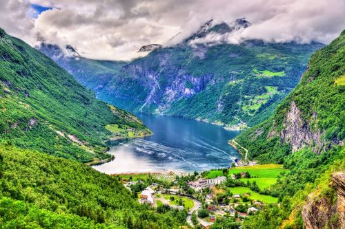 A deep blue fjord winds between steep green mountains with scattered villages, farmland, and low clouds hanging over the peaks.